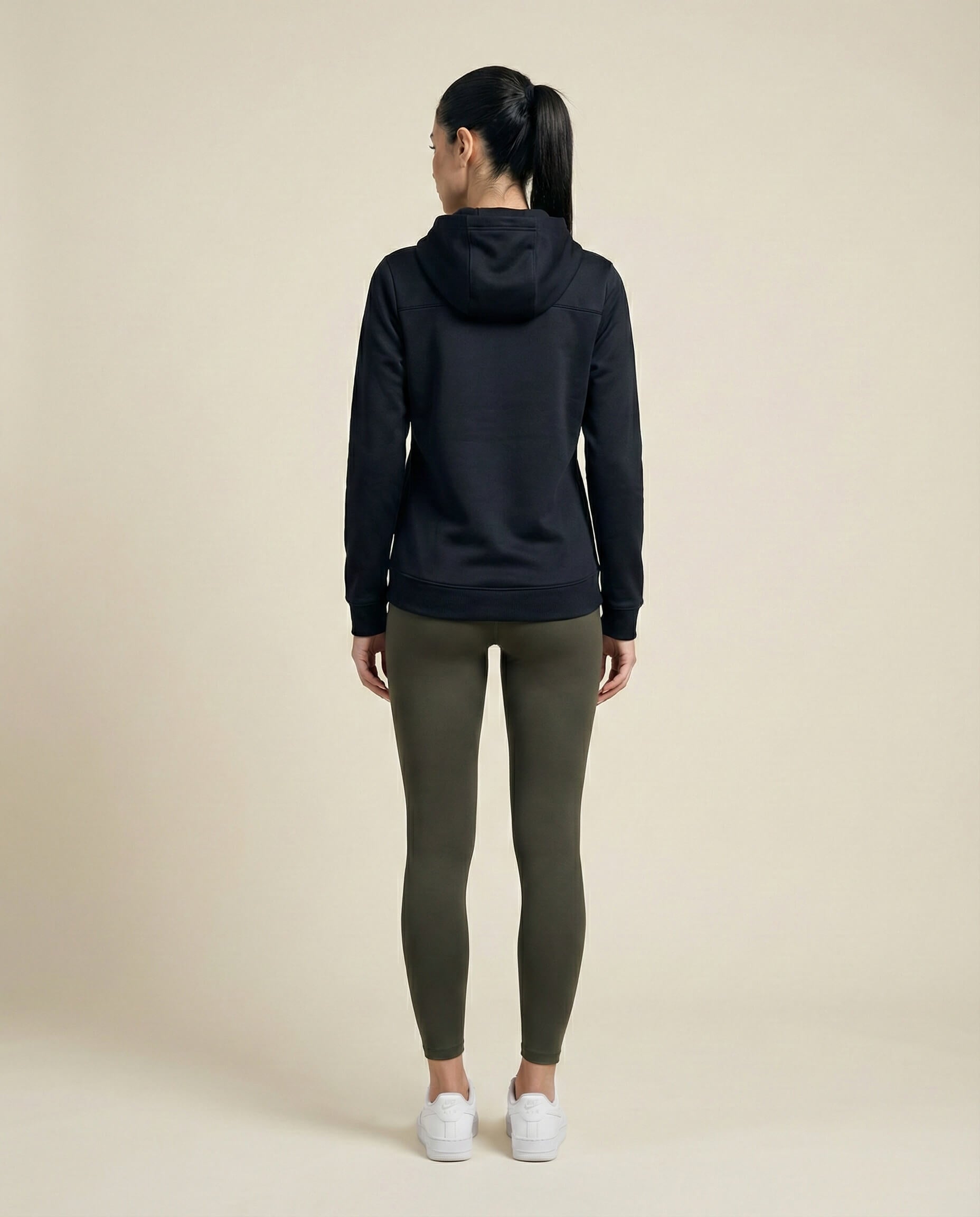 Woman in black hoodie and olive leggings, white sneakers, standing against neutral background