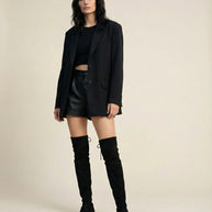 Woman in black blazer, crop top, leather shorts, and knee-high boots standing indoors