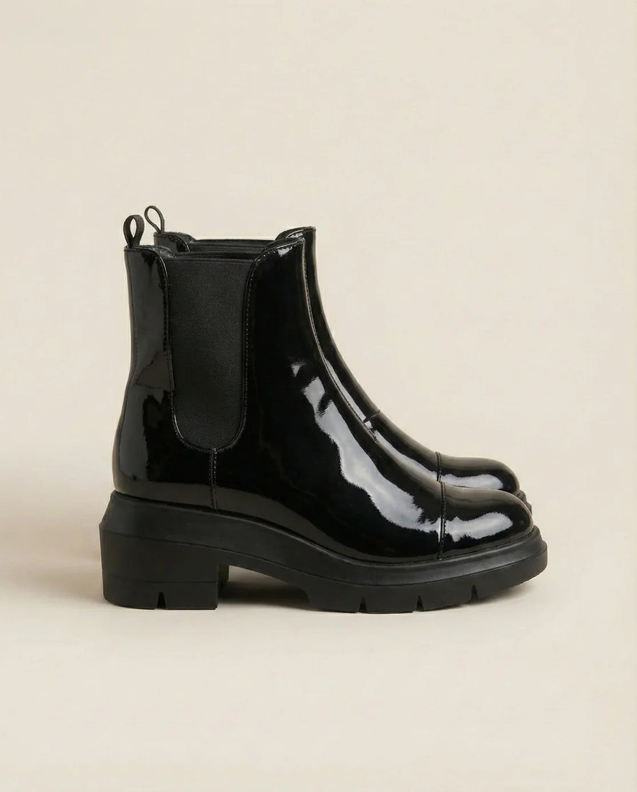 Shiny black patent leather Chelsea boots with chunky rubber soles on a beige background
