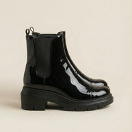Shiny black patent leather Chelsea boots with chunky rubber soles on a beige background