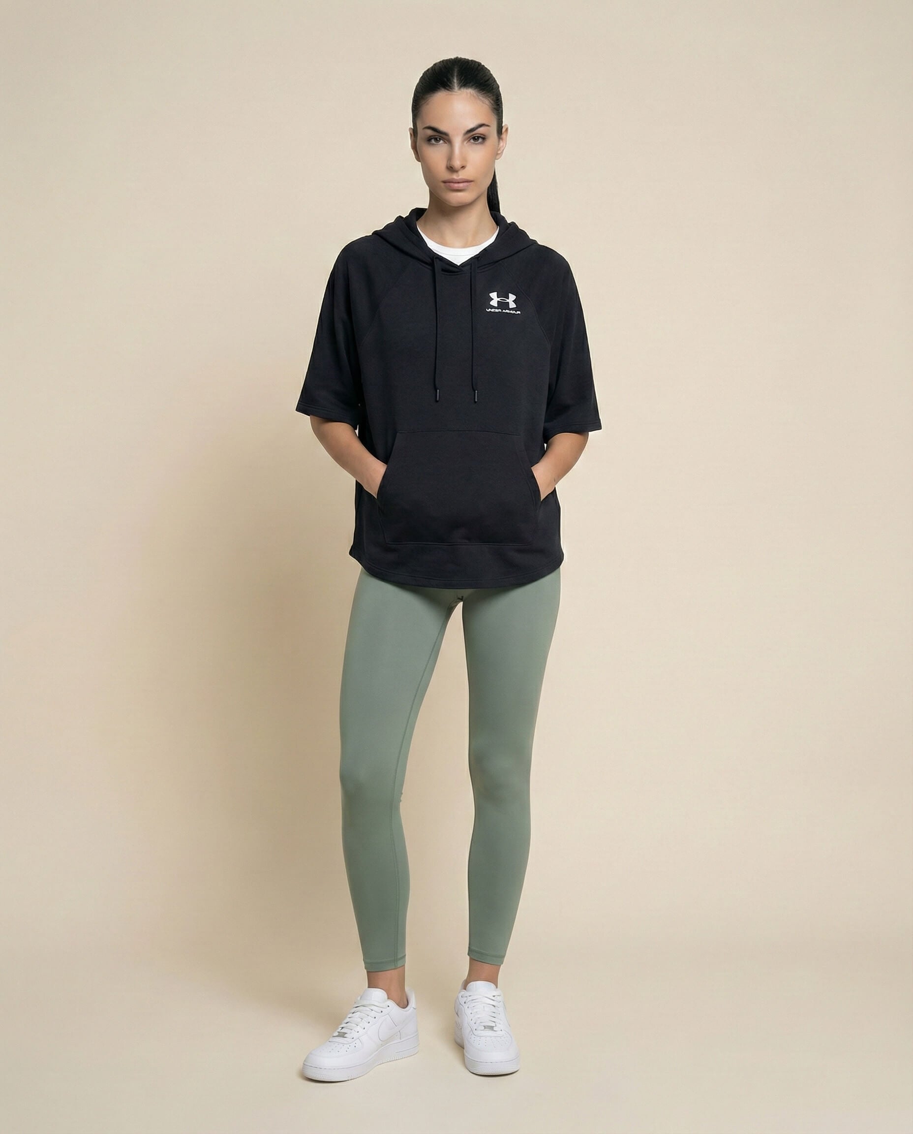 Woman in black Under Armour hoodie, sage green leggings, and white sneakers standing against beige background