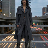Woman wearing gray trench coat and boots standing on city overpass at dusk