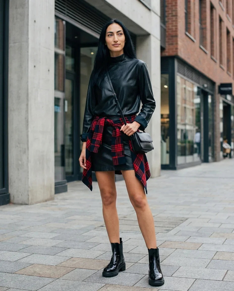 Woman in black leather top and skirt, plaid shirt tied at waist, black boots, city street style