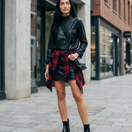 Woman in black leather top and skirt, plaid shirt tied at waist, black boots, city street style