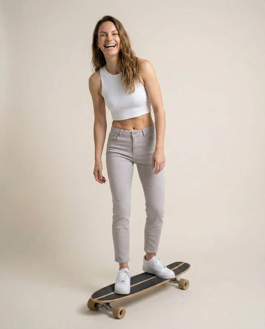 Smiling woman in white crop top and light jeans standing on skateboard indoors