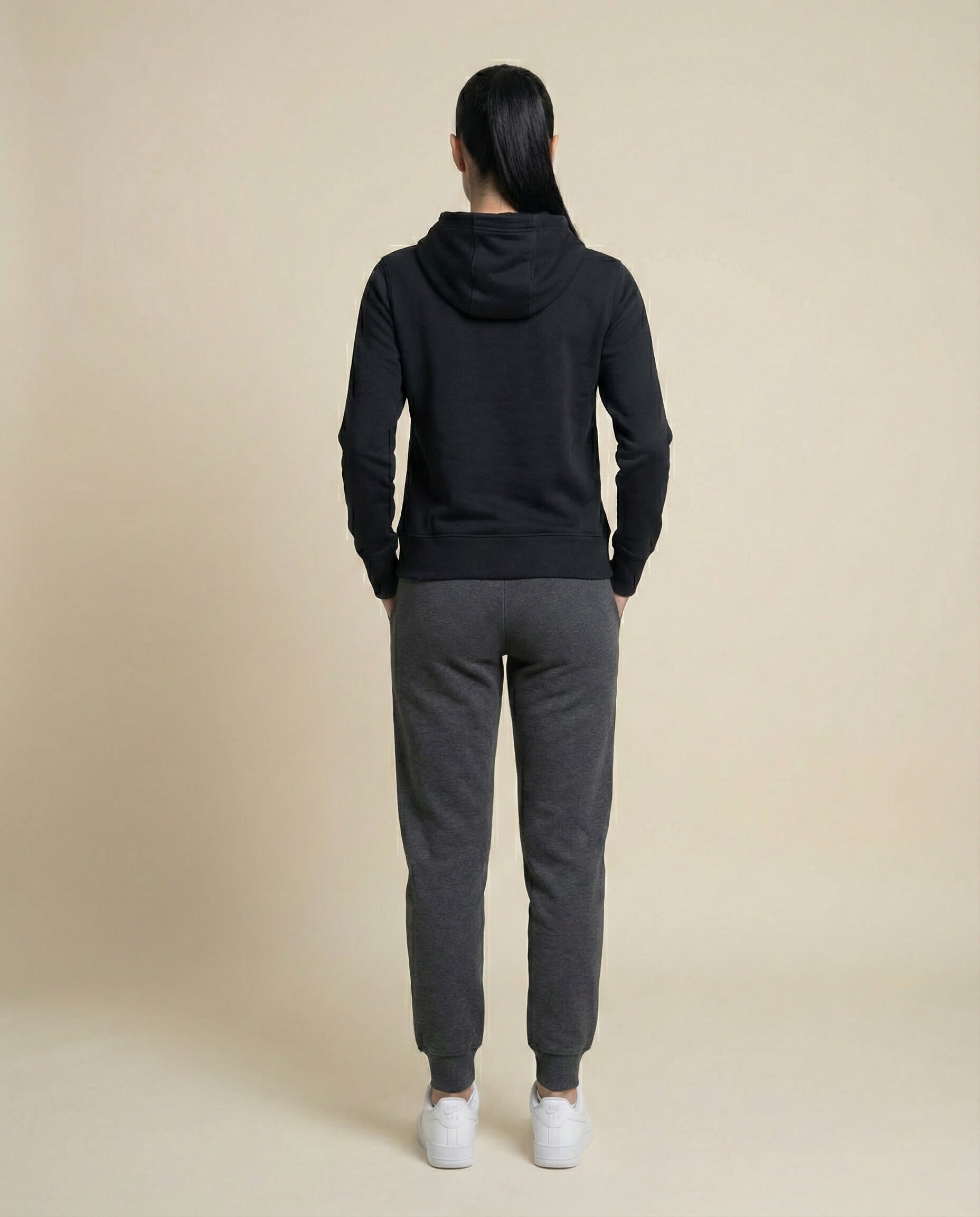 Person in black hoodie and gray sweatpants with white sneakers, standing on beige background