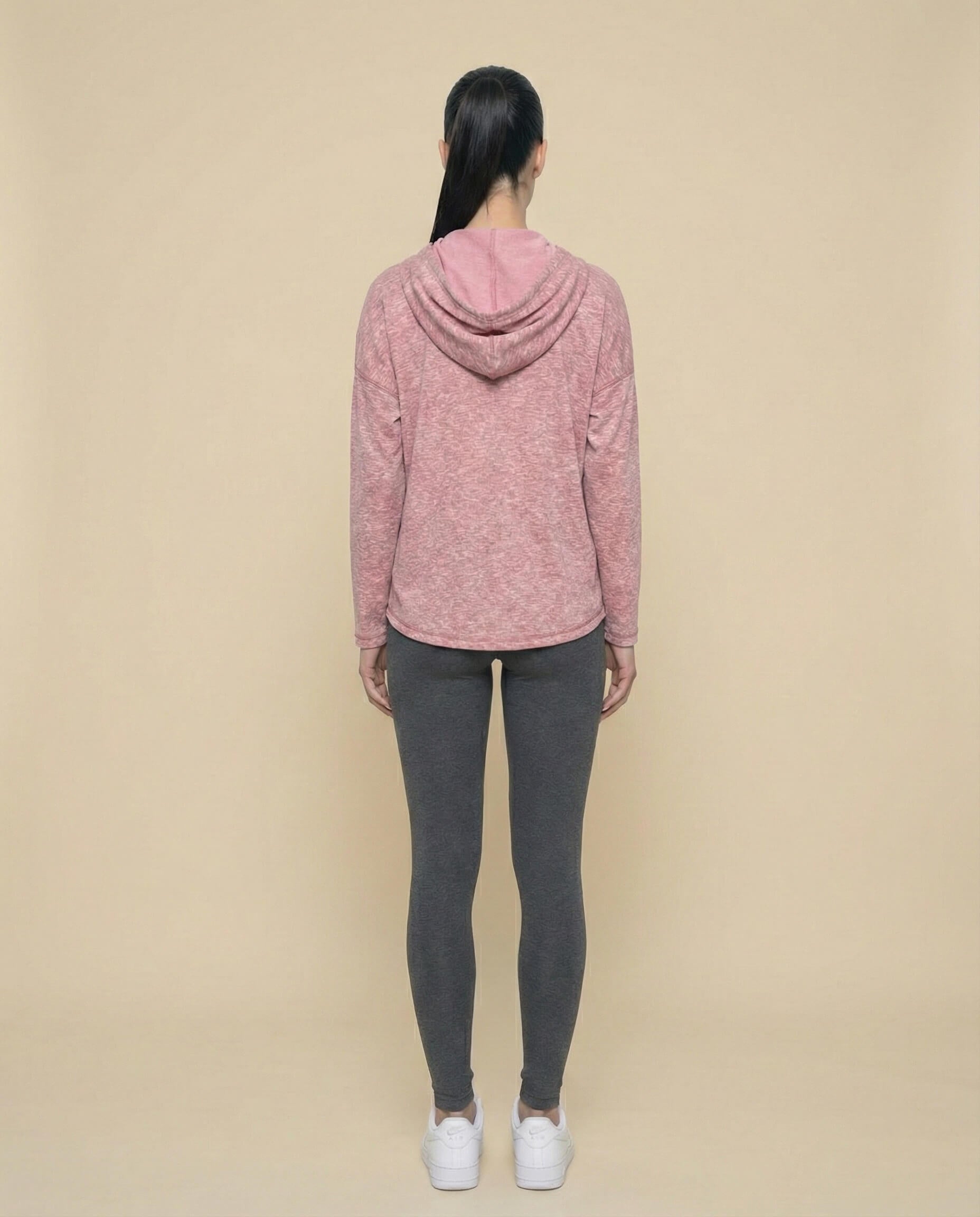 Woman in pink hooded sweatshirt, gray leggings, and white sneakers, standing against beige background