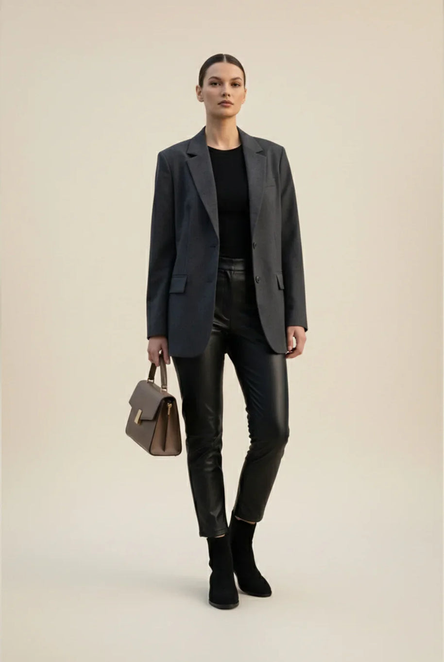Woman in gray blazer, black top, leather pants, ankle boots, carrying brown handbag
