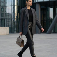 Woman in black blazer and leather pants walking outside modern glass building, holding handbag