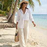 Woman in white linen outfit, straw hat, and sunglasses walking on tropical beach with palm trees