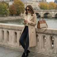 Woman in beige trench coat, turtleneck sweater, and sunglasses standing by riverside with handbag