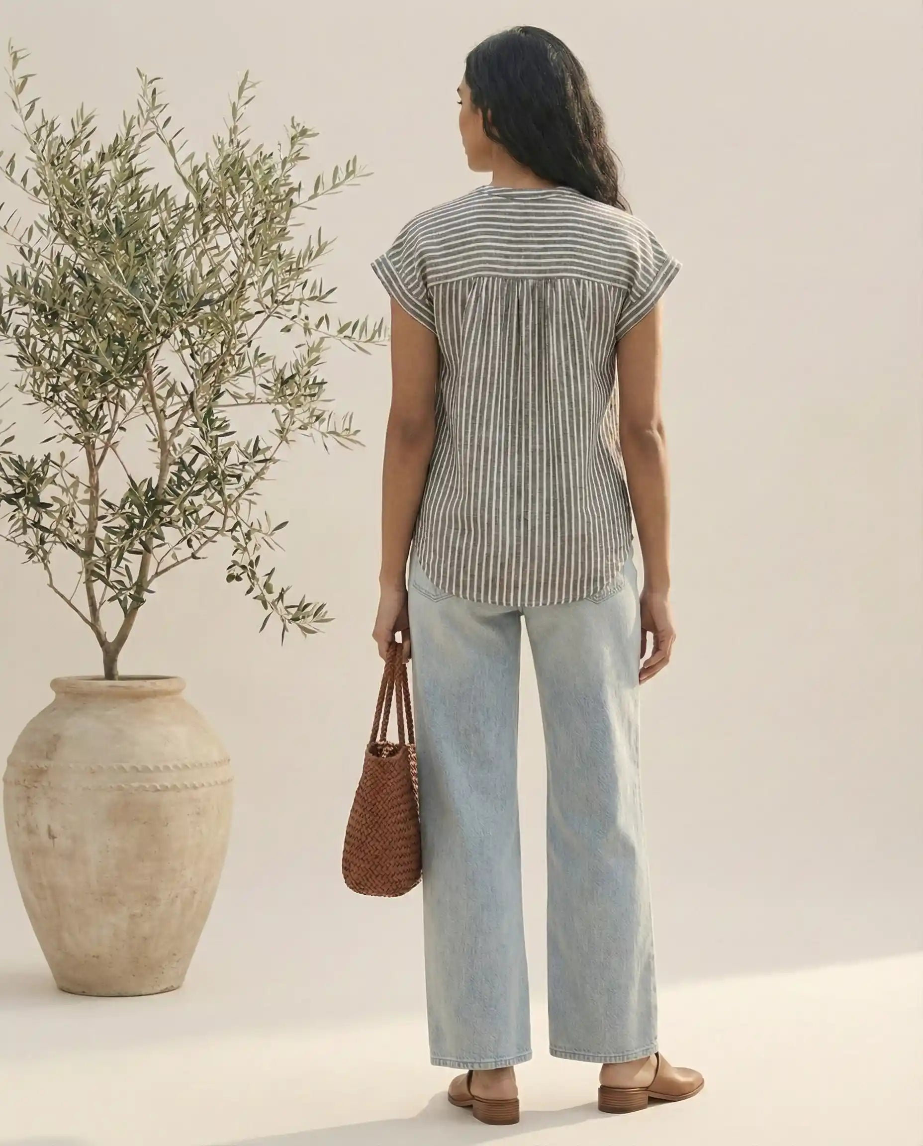 Woman in striped short-sleeve blouse and light blue jeans holding woven bag, standing by potted plant