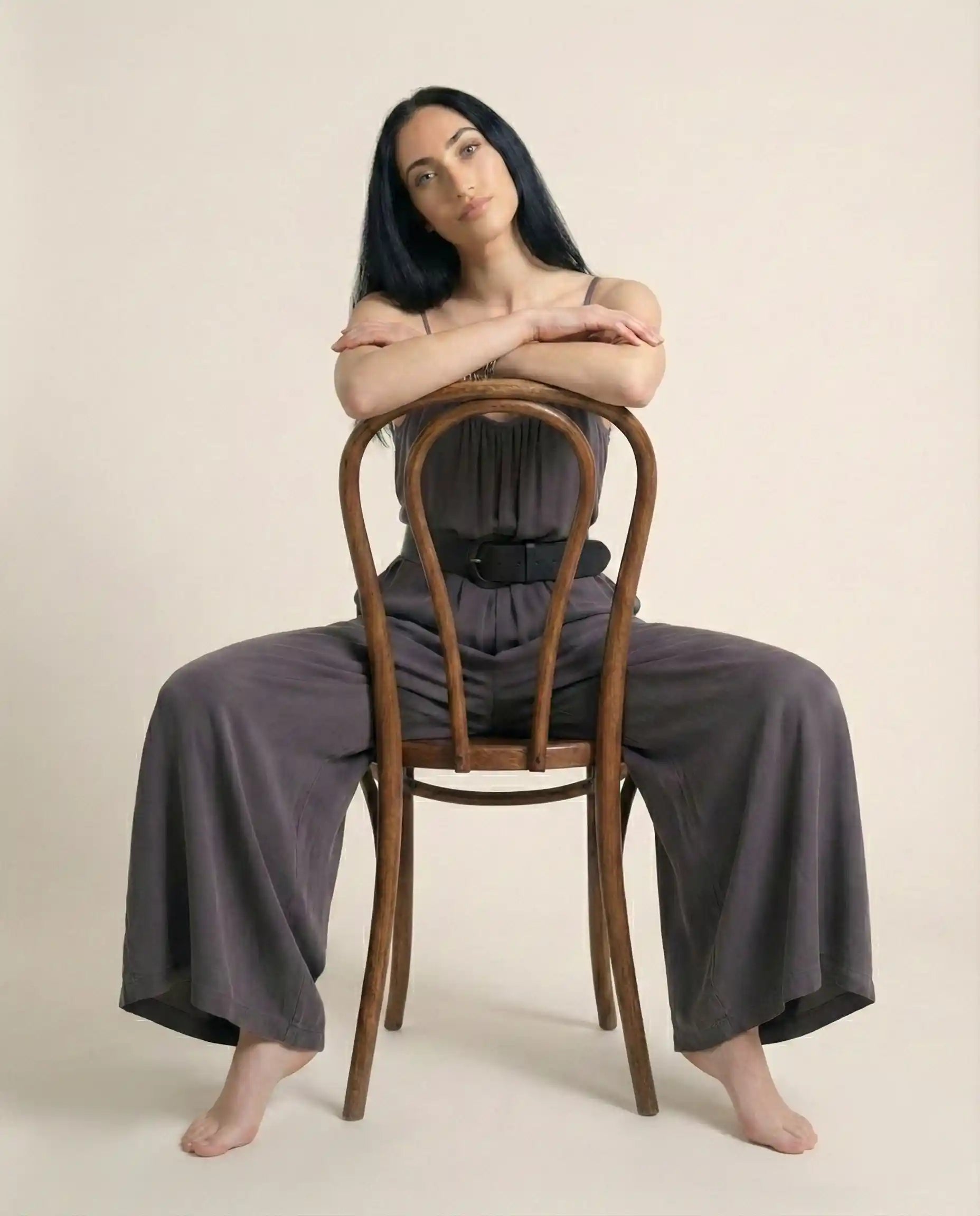 Woman in gray wide-leg jumpsuit sitting backwards on a wooden chair on beige background