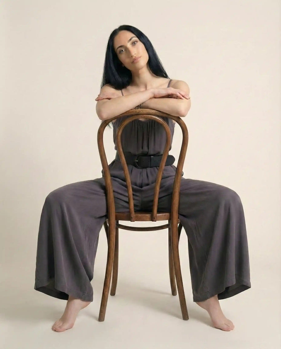 Woman in gray wide-leg jumpsuit sitting backwards on a wooden chair on beige background