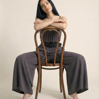 Woman in gray wide-leg jumpsuit sitting backwards on a wooden chair on beige background
