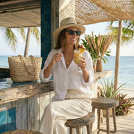 Woman in sun hat and sunglasses relaxing at beach bar, holding tropical drink, straw bag on counter