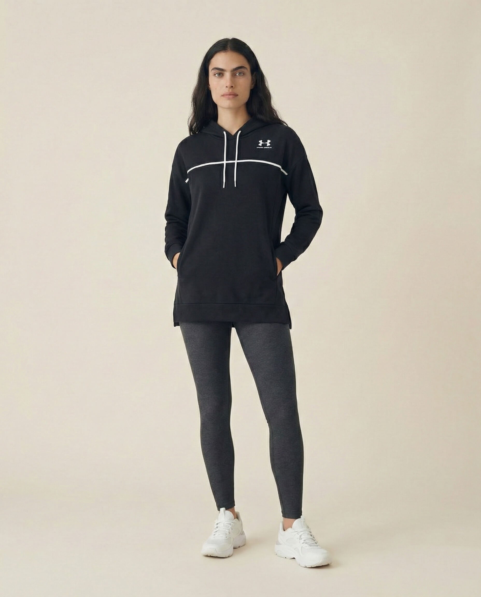 Woman wearing a black athletic hoodie, gray leggings, and white sneakers against plain background