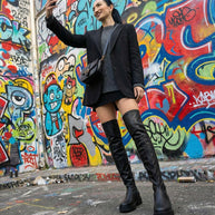 Woman taking selfie in front of vibrant graffiti wall, wearing black blazer and thigh-high boots