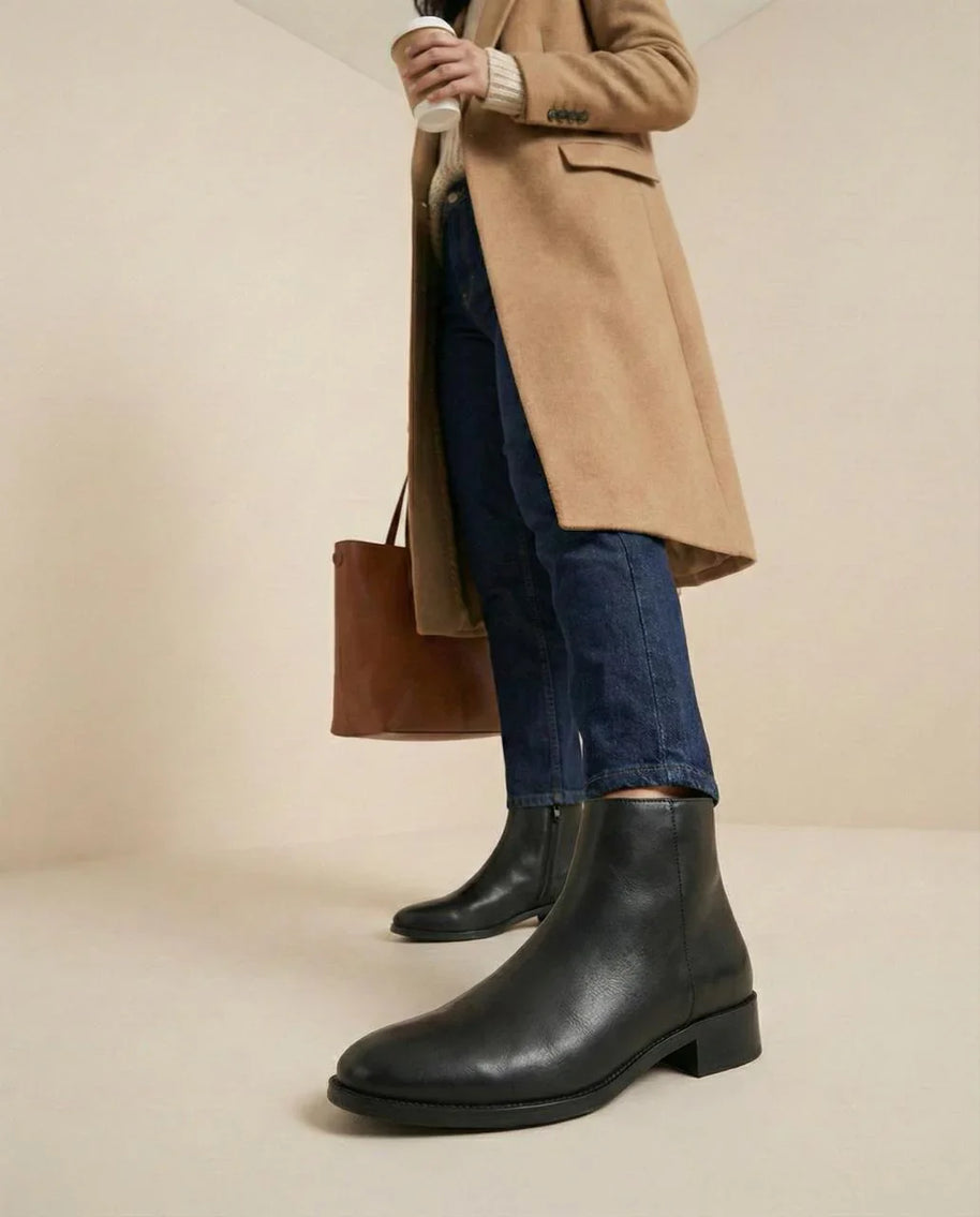 Woman wearing black leather ankle boots, blue jeans, camel coat, holding coffee cup