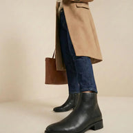 Woman wearing black leather ankle boots, blue jeans, camel coat, holding coffee cup