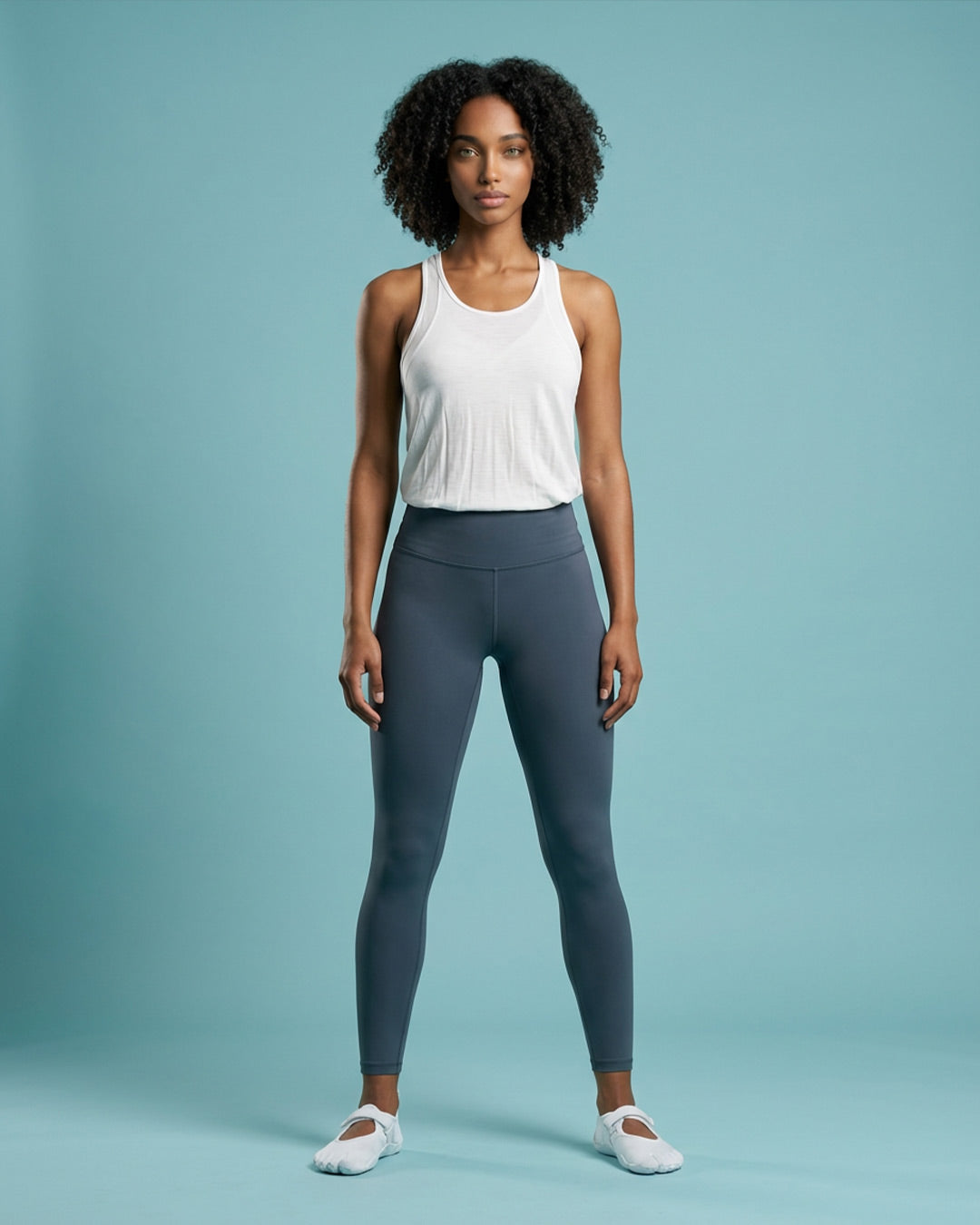 Woman in white sleeveless athletic top and gray leggings standing against a blue background