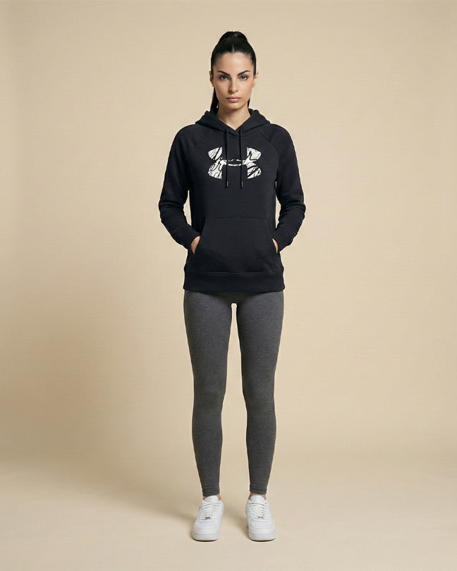 Woman in black graphic hoodie, gray leggings, and white sneakers standing against beige background.