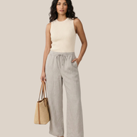 Woman in sleeveless knit top and linen pants holding straw tote bag, wearing sandals