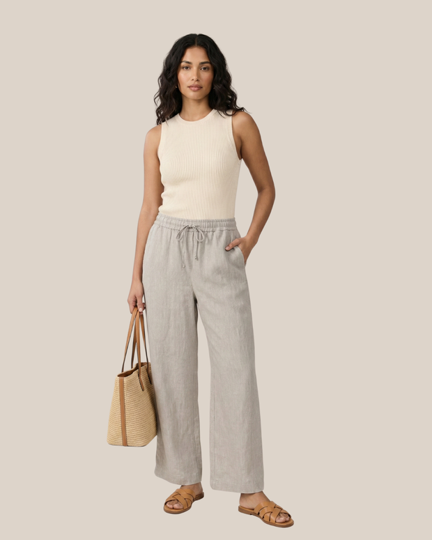 Woman in sleeveless knit top and linen pants holding straw tote bag, wearing sandals