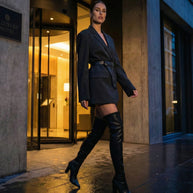Woman in a belted blazer and thigh-high black boots standing outside a luxury hotel entrance.
