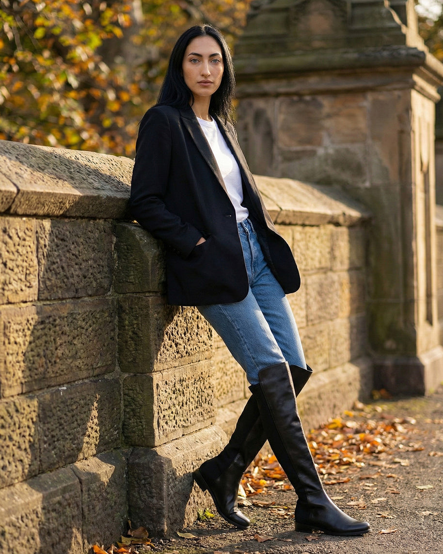 Woman in black blazer, jeans, and knee-high black boots leaning against stone wall outdoors