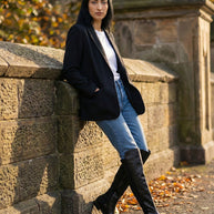 Woman in black blazer, jeans, and knee-high black boots leaning against stone wall outdoors