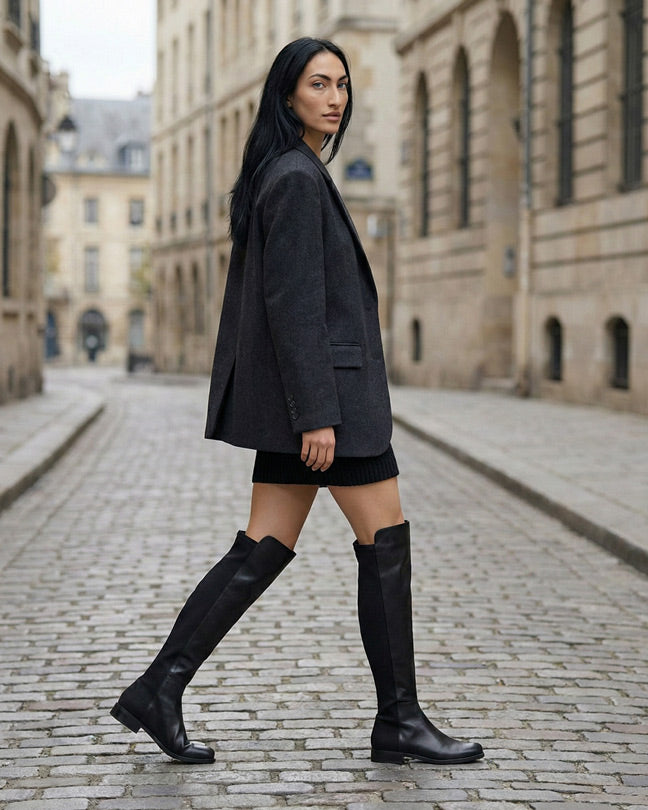 Woman in black knee-high boots, black skirt, and gray blazer walking on a cobblestone street