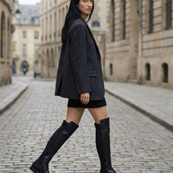Woman in black knee-high boots, black skirt, and gray blazer walking on a cobblestone street