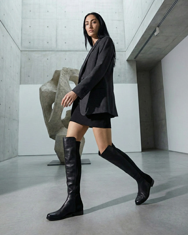 Woman in black blazer and mini skirt wearing tall black boots in a modern gallery setting