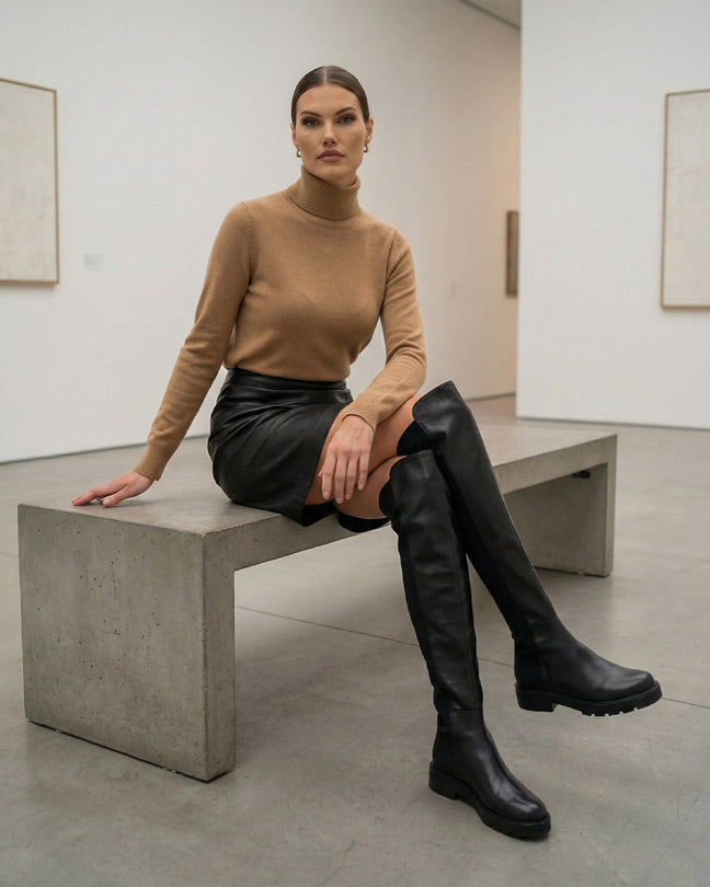 Woman in a camel turtleneck, black leather mini skirt, and over-the-knee black boots sitting on a concrete bench in a modern art gallery.