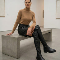 Woman in a camel turtleneck, black leather mini skirt, and over-the-knee black boots sitting on a concrete bench in a modern art gallery.