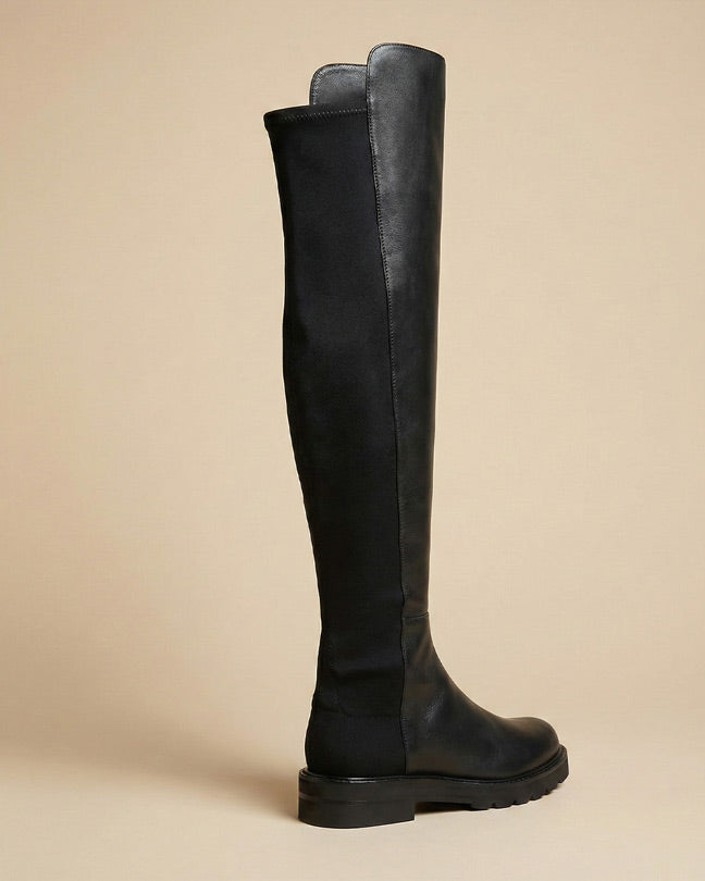 Black over-the-knee boot with flat sole, leather and suede design, side profile on beige background