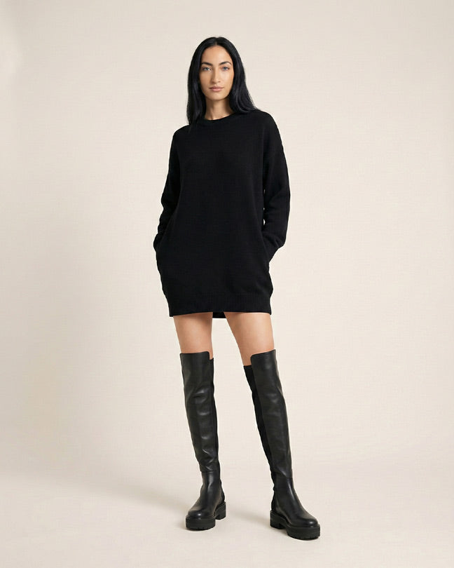 Woman wearing a black sweater dress and black knee-high boots, standing against a plain background.