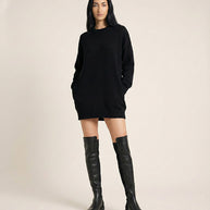 Woman wearing a black sweater dress and black knee-high boots, standing against a plain background.