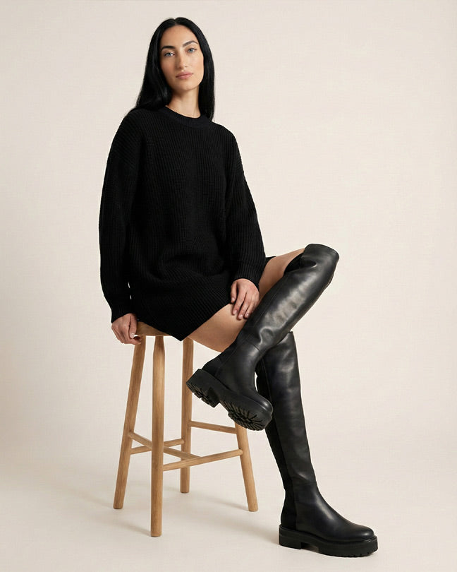 Woman in black sweater dress and black over-the-knee boots sitting on wooden stool