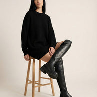 Woman in black sweater dress and black over-the-knee boots sitting on wooden stool