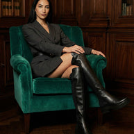 Woman in blazer and black knee-high boots seated on green armchair in vintage library