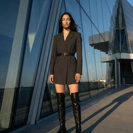 Woman in belted black blazer dress and knee-high boots standing by modern glass building