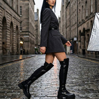 Woman wearing a black blazer dress and knee-high black boots walking on a cobblestone street