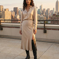 Woman in a beige satin shirt dress with side slit and black knee-high boots on a rooftop