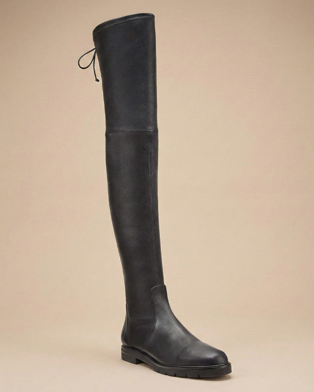 Black over-the-knee leather boot with low heel and tie detail, side view on beige background