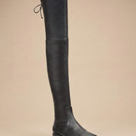 Black over-the-knee leather boot with low heel and tie detail, side view on beige background
