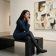 Woman in denim outfit and black knee-high boots sitting on bench in modern art gallery