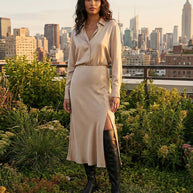 Woman in a beige satin shirt dress with side slit and black knee-high boots on rooftop garden