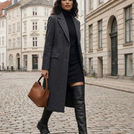 Woman in a grey coat, black dress, and knee-high boots walking with a brown handbag on a city street